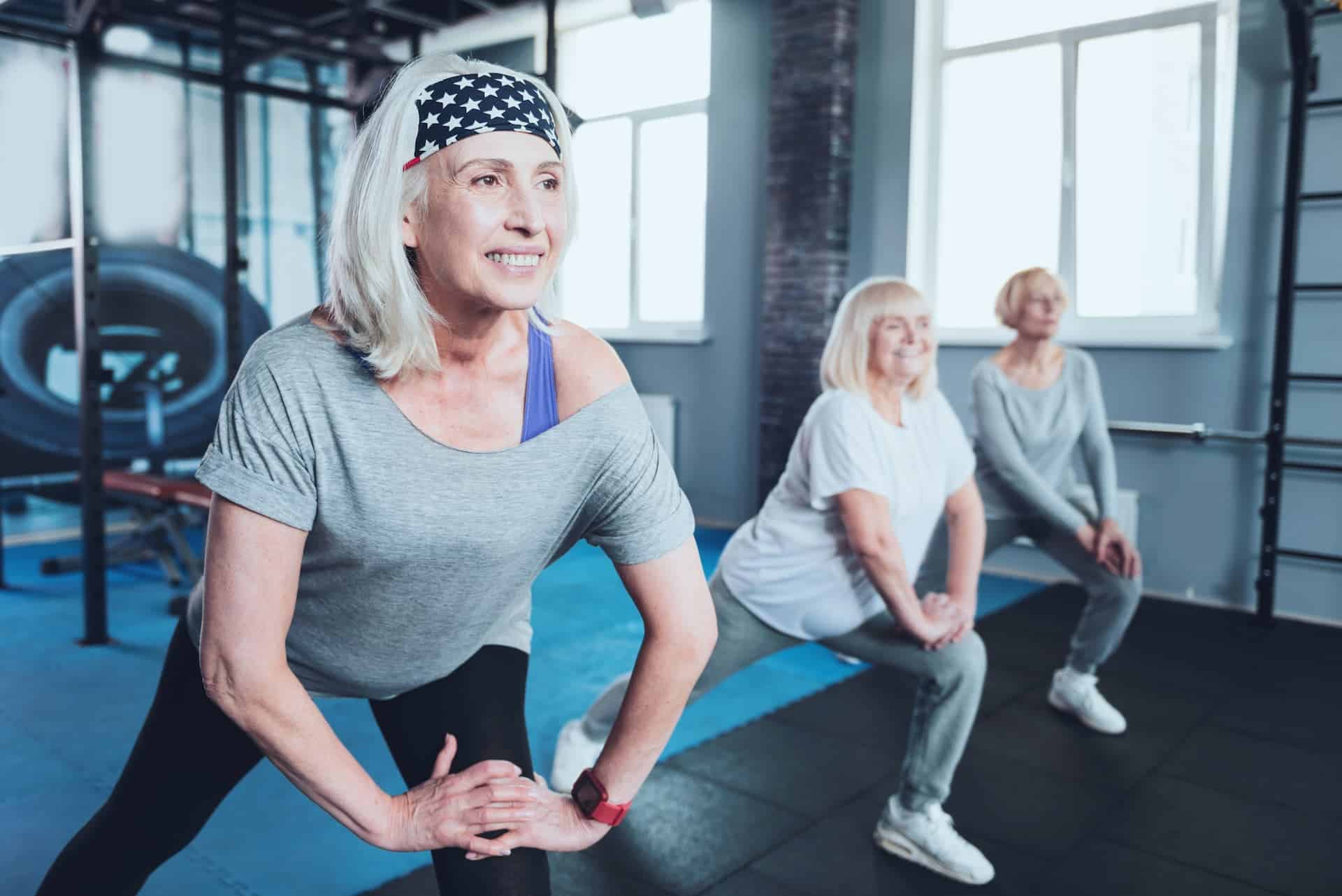 Selective focus on a joyful woman smiling while lunging with a group of elderly ladies while all taking an exercise class at a fitness club after conquering knee pain.