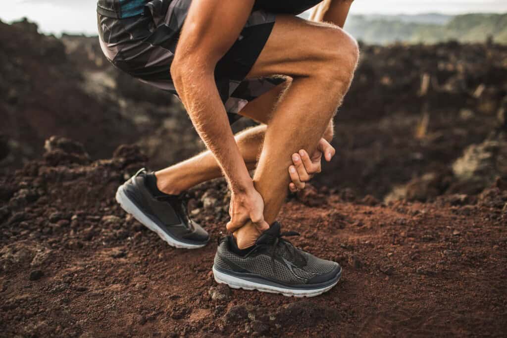 Close-up of a male runner's legs and feet, bent over and clutching his Achilles tendon in pain.