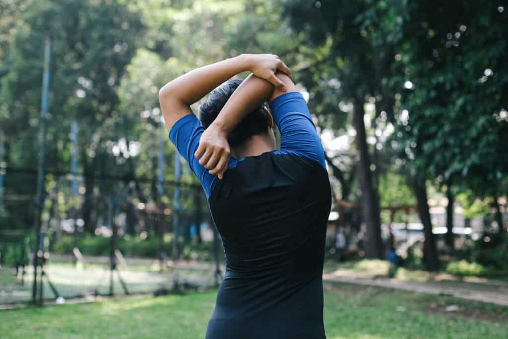 A man wearing a blue and black shirt, doing elbow stretches by stretching his arms over his head and behind his back in a park with trees and grass in the background. 