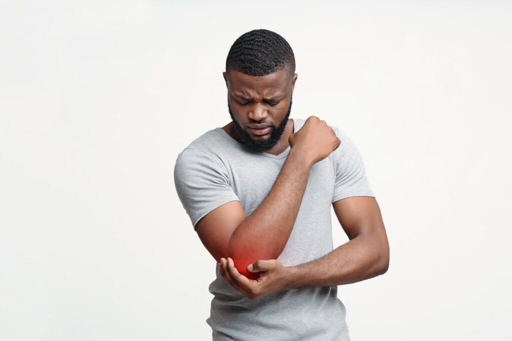 A man in a gray shirt holds his left elbow, which appears to be in pain from tennis elbow, with his right hand.