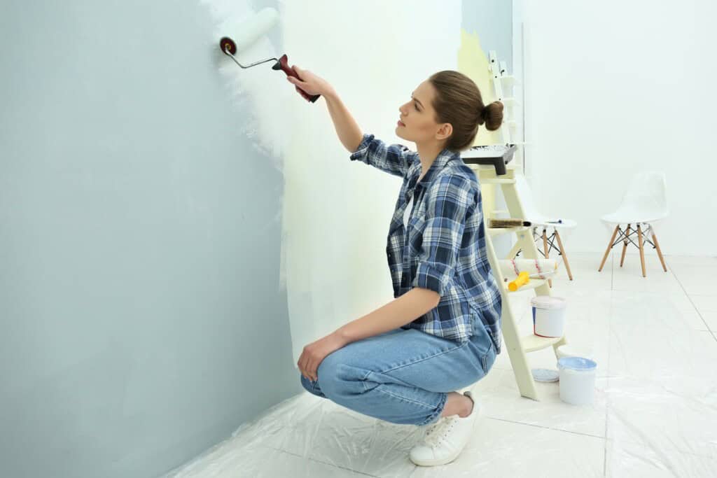 A woman kneeling while painting a wall in her home.