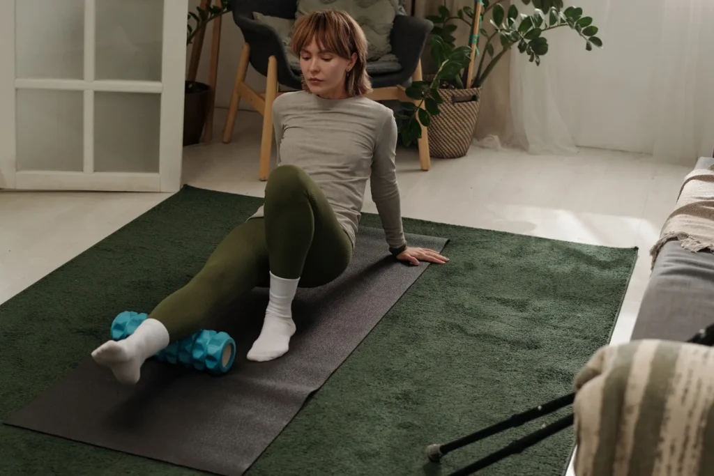 Woman using a foam roller on her calf while sitting on a yoga mat in a cozy home setting, practicing self-massage for muscle relief.