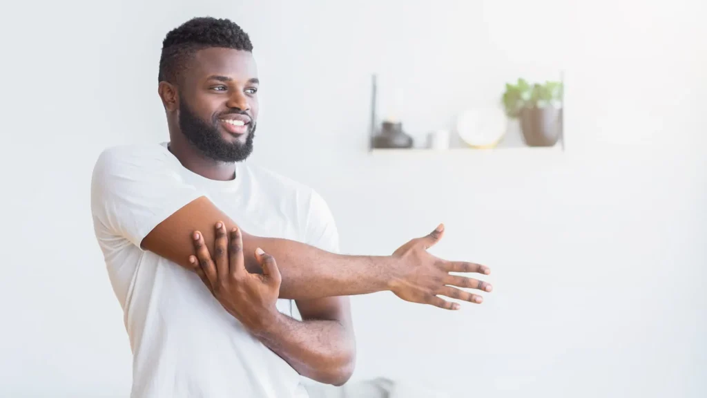 Smiling man in a white t-shirt gently stretching his shoulder and arm across his chest at home, demonstrating a mobility exercise often used in rotator cuff injury recovery and shoulder self-care.