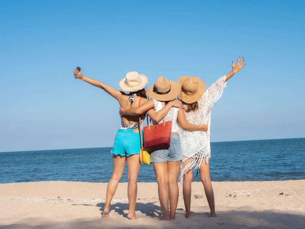 Rear view of three women standing arm-in-arm at the beach, looking out at the ocean, confidently showing their legs and enjoying the sun after bodywork for cellulite reduction.