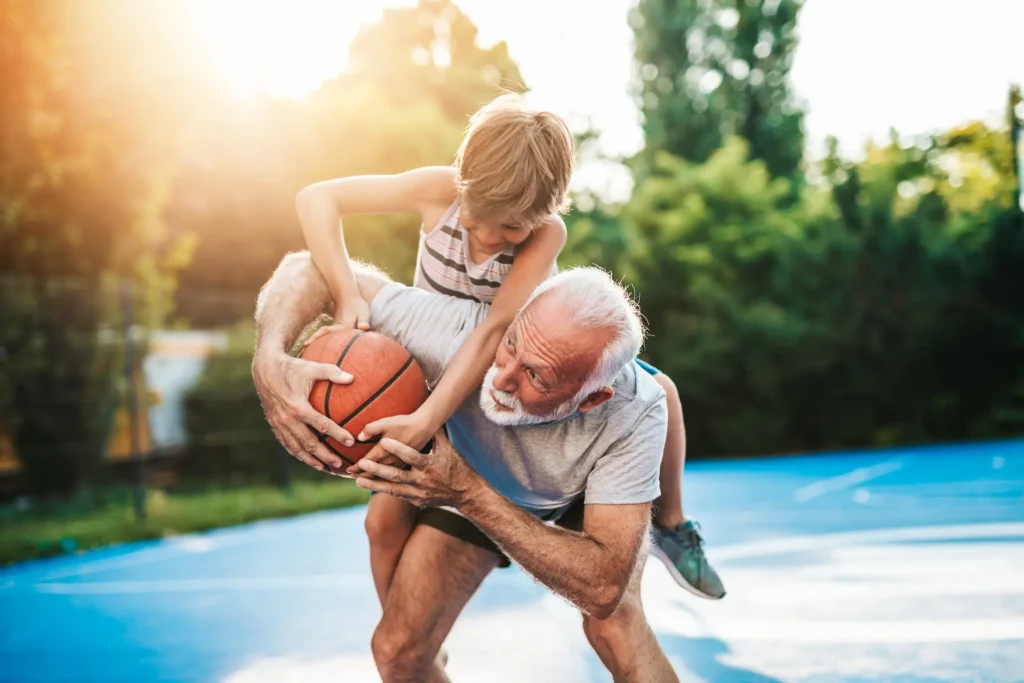 A grandfather plays basketball with his grandson outdoors. The boy is on his back, reaching playfully for the ball as they laugh and enjoy a sunny day together.