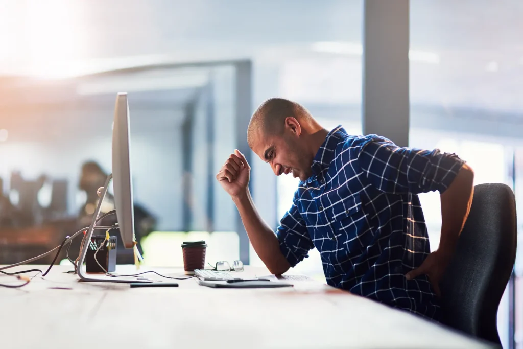 Man at desk holding his lower back in discomfort, showing signs of L5-S1 strain or lumbosacral dysfunction—common issues that can benefit from targeted bodywork or massage therapy.