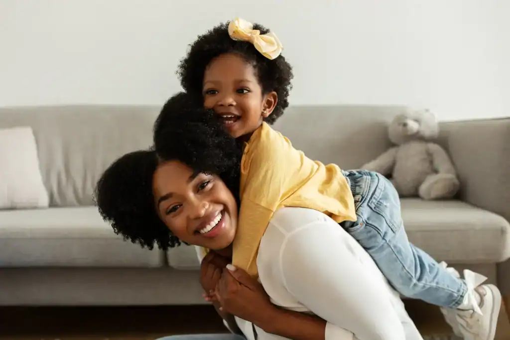 Mother smiling as she crouches playfully with her young daughter on her back, both laughing and enjoying the moment—illustrating the freedom of movement and connection made possible after bodywork for Sacroiliac Joint Pain.