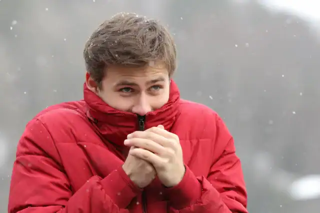 Man in a red puffer jacket warming his hands near his face on a cold day, a common response to the cold that can leave shoulders and neck tense.
