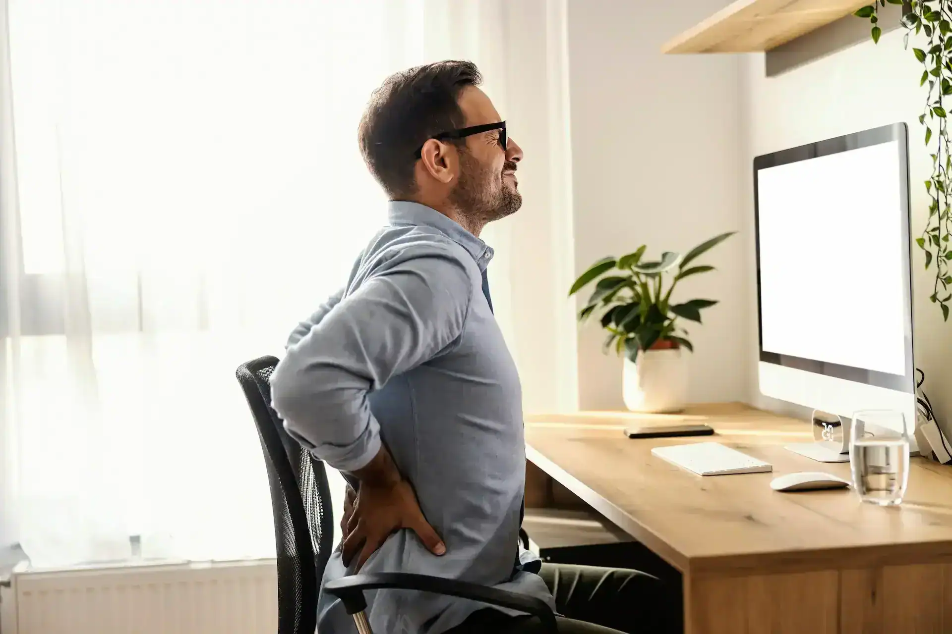 Man sitting at a desk holding his lower back in discomfort, showing typical posture-related Sacroiliac Joint Pain often relieved by bodywork and massage.