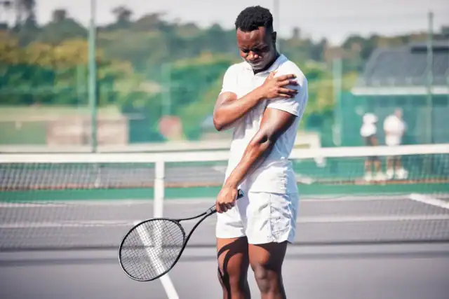 Tennis player standing on the court holding his shoulder while gripping a racket, showing discomfort during play.