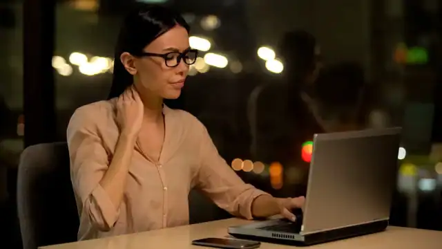 Woman touching her neck while working on a laptop, illustrating end-of-day discomfort from prolonged posture.